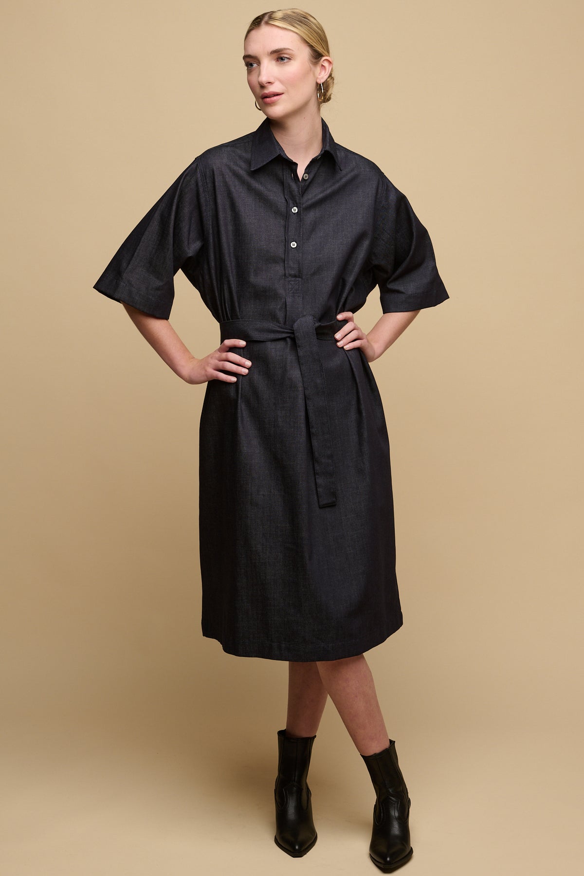Full body image of blonde female wearing collared straight denim dress in indigo with 3/4 sleeves and waist belt tie, female holding hands on hips. Worn with heeled black boots.