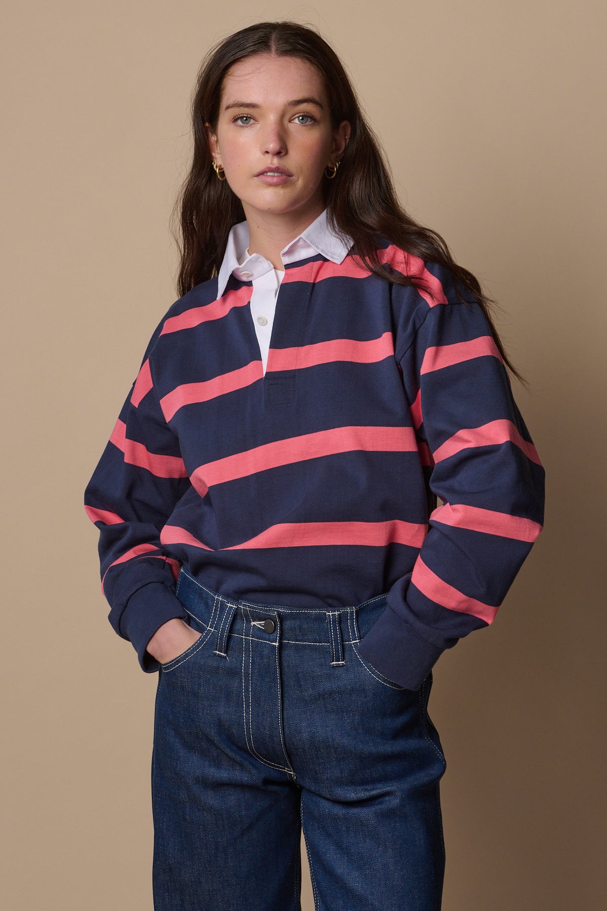 Thigh up image of white female wearing unisex striped rugby shirt in navy pink  tucked into jeans 