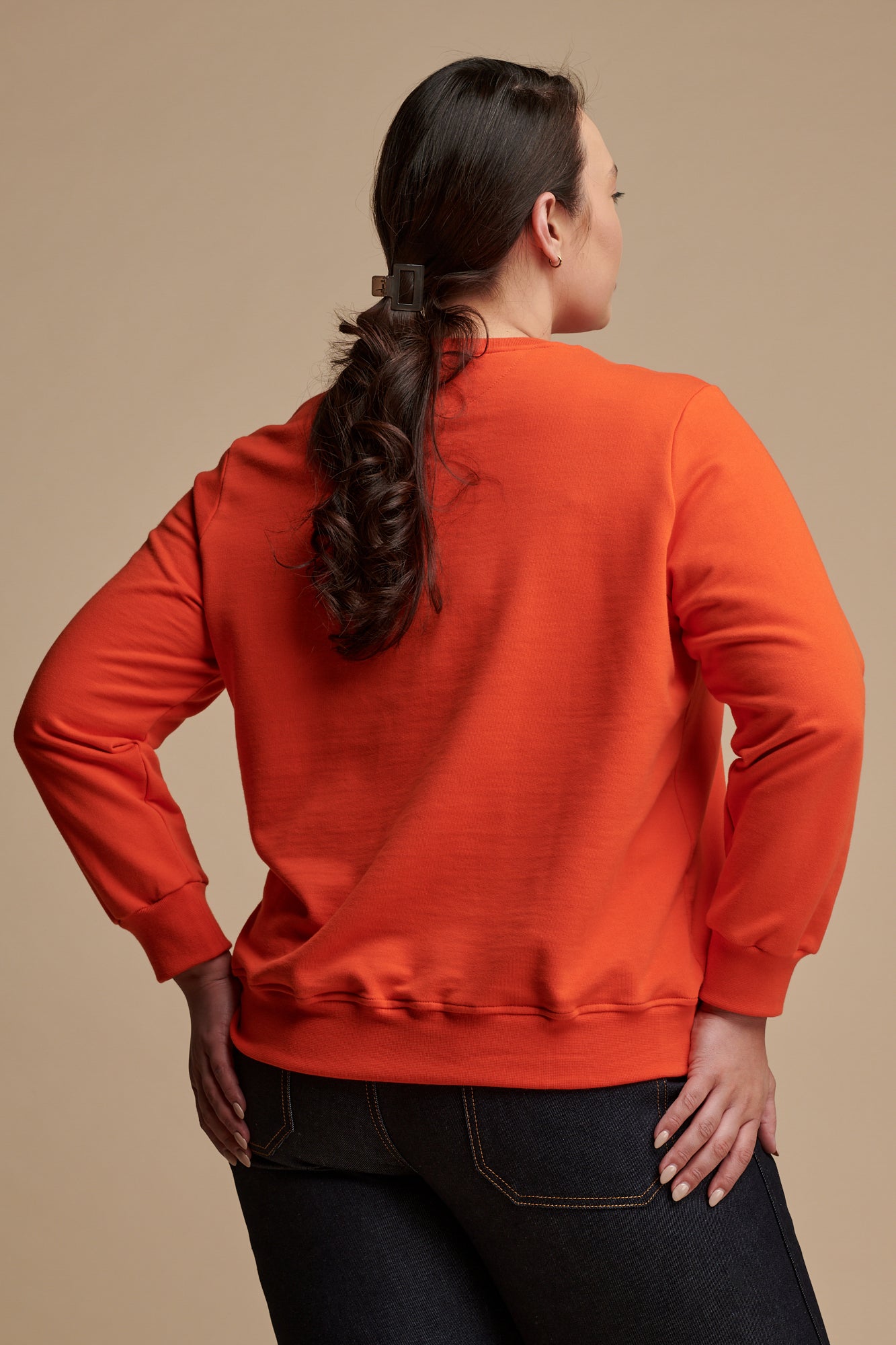 Thigh up of smiley female with dark hair tied back wearing sweatshirt in flame red  paired with work jeans in indigo