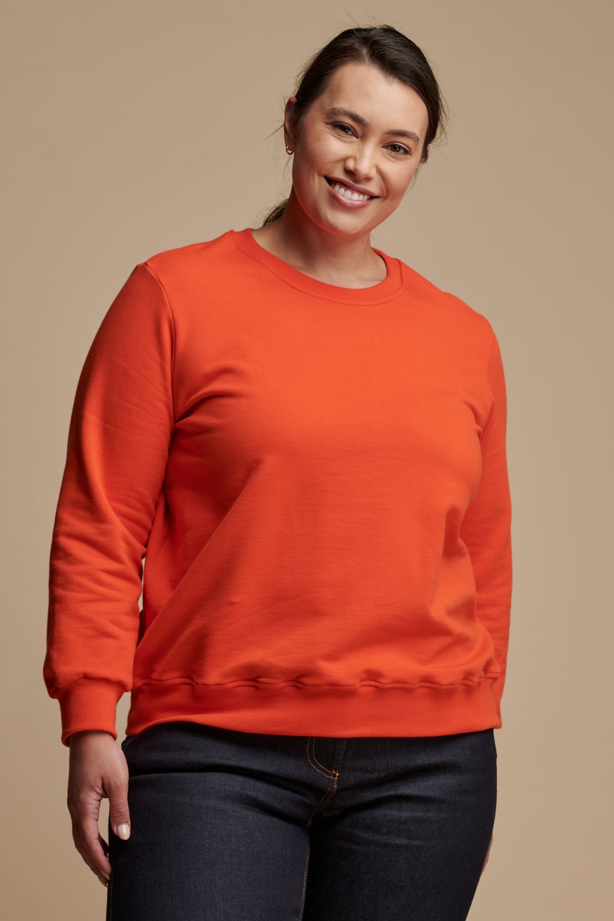 Thigh up of smiley female with dark hair tied back wearing sweatshirt in flame red  paired with work jeans in indigo