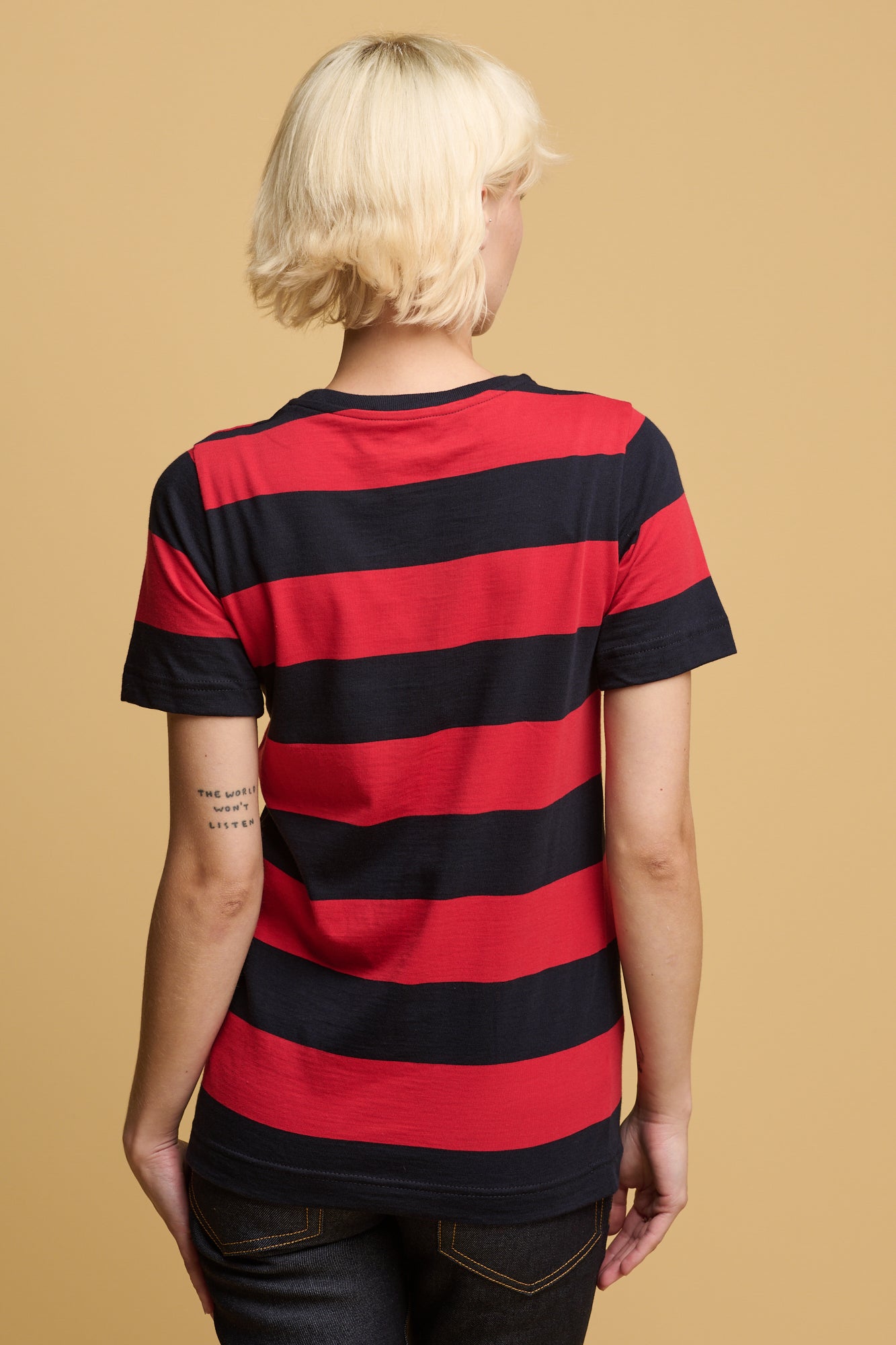 Blonde female tood with one leg infront of the other with her arms resting behind her waist wearing wide stripe short sleeve t shirt in navy and red paired with straight leg jeans in indigo