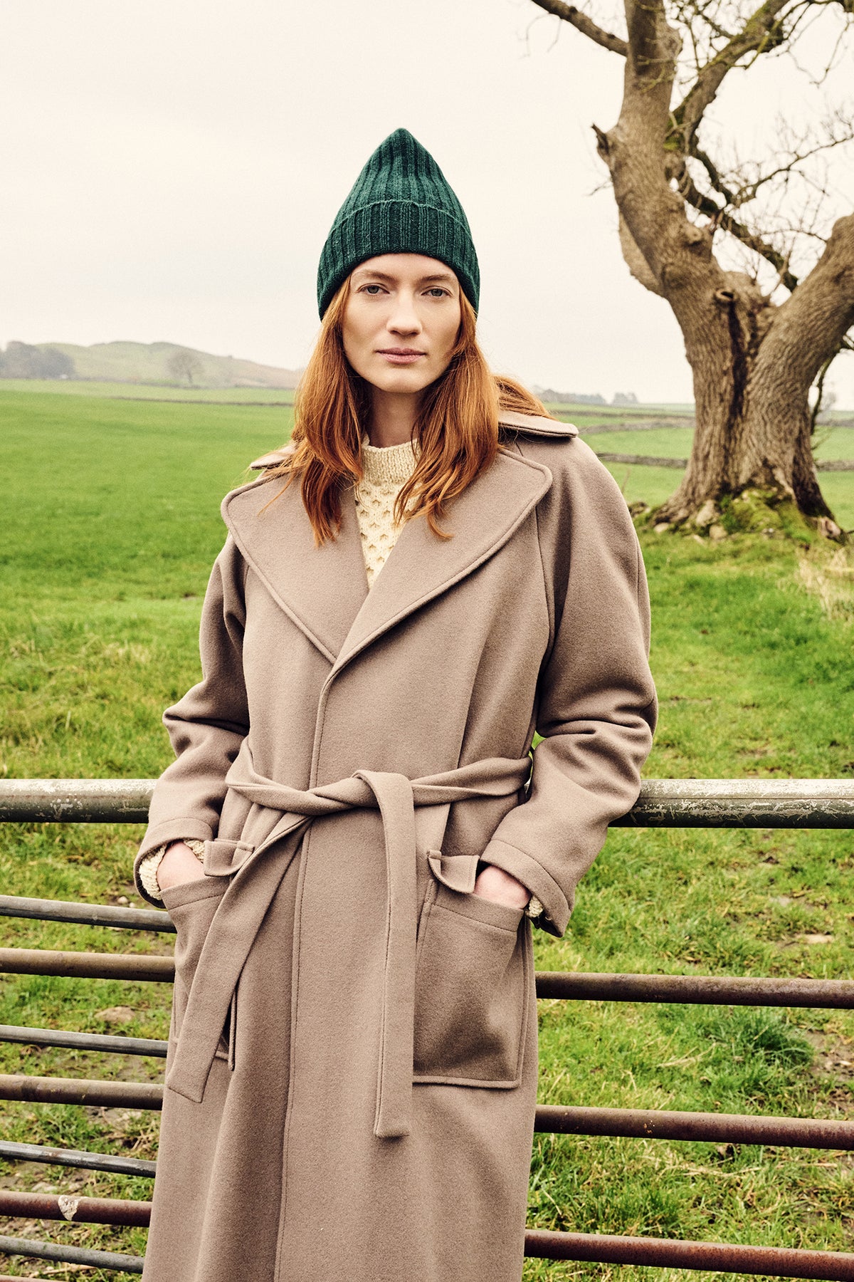Woman wearing a long wool coat and green beanie standing in a field with a tree in the background