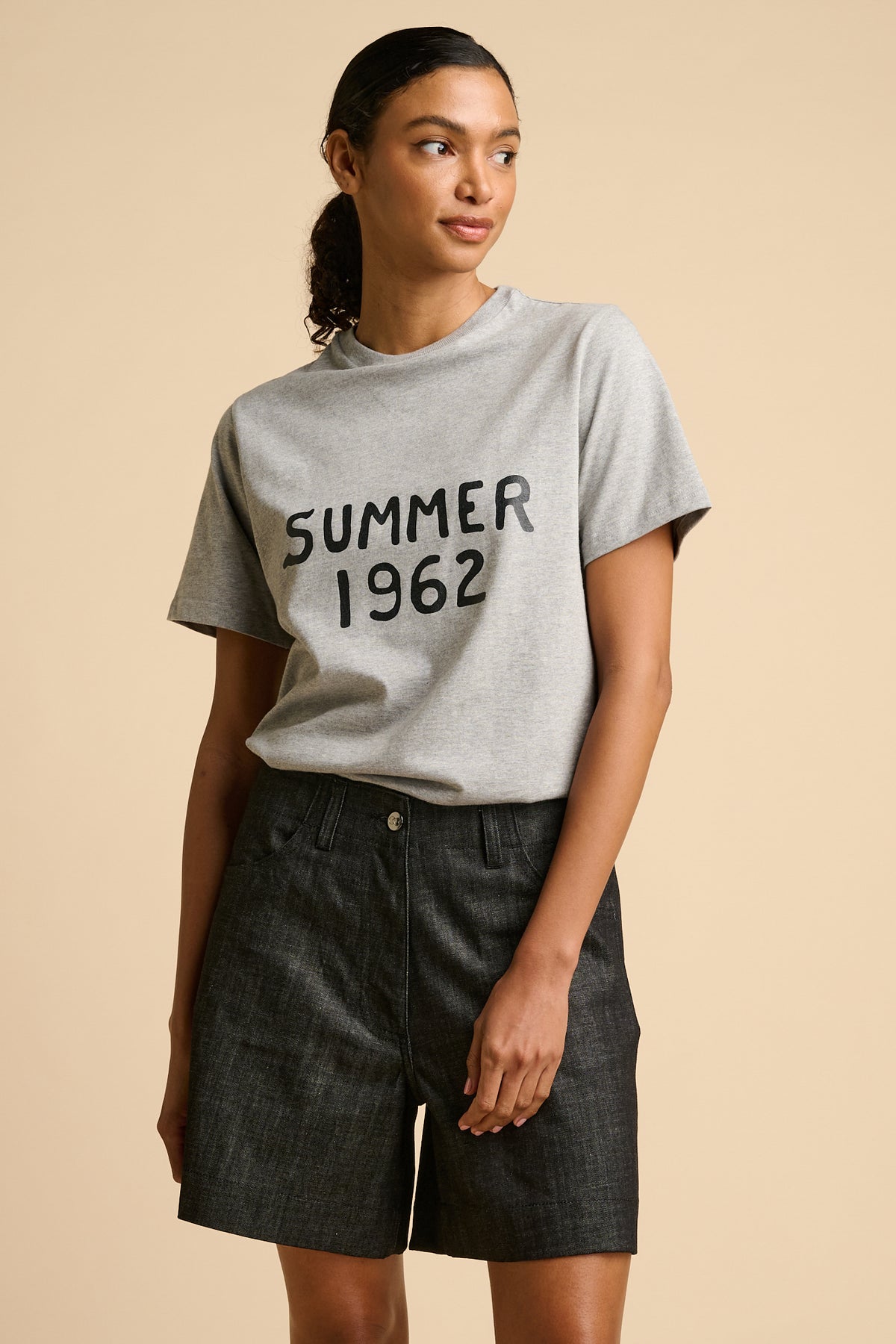 Woman wearing a grey t-shirt with 'Summer 1962' text with black shorts on a beige background.