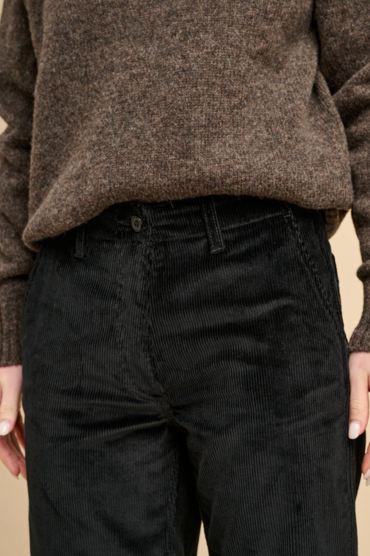 Person wearing a brown sweater and black corduroy pants against a beige background
