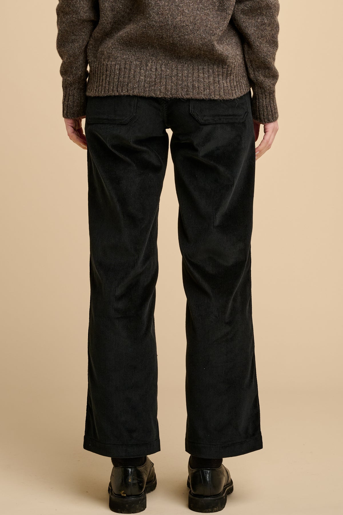 The back of woman woman wearing black corduroy trousers against beige background.