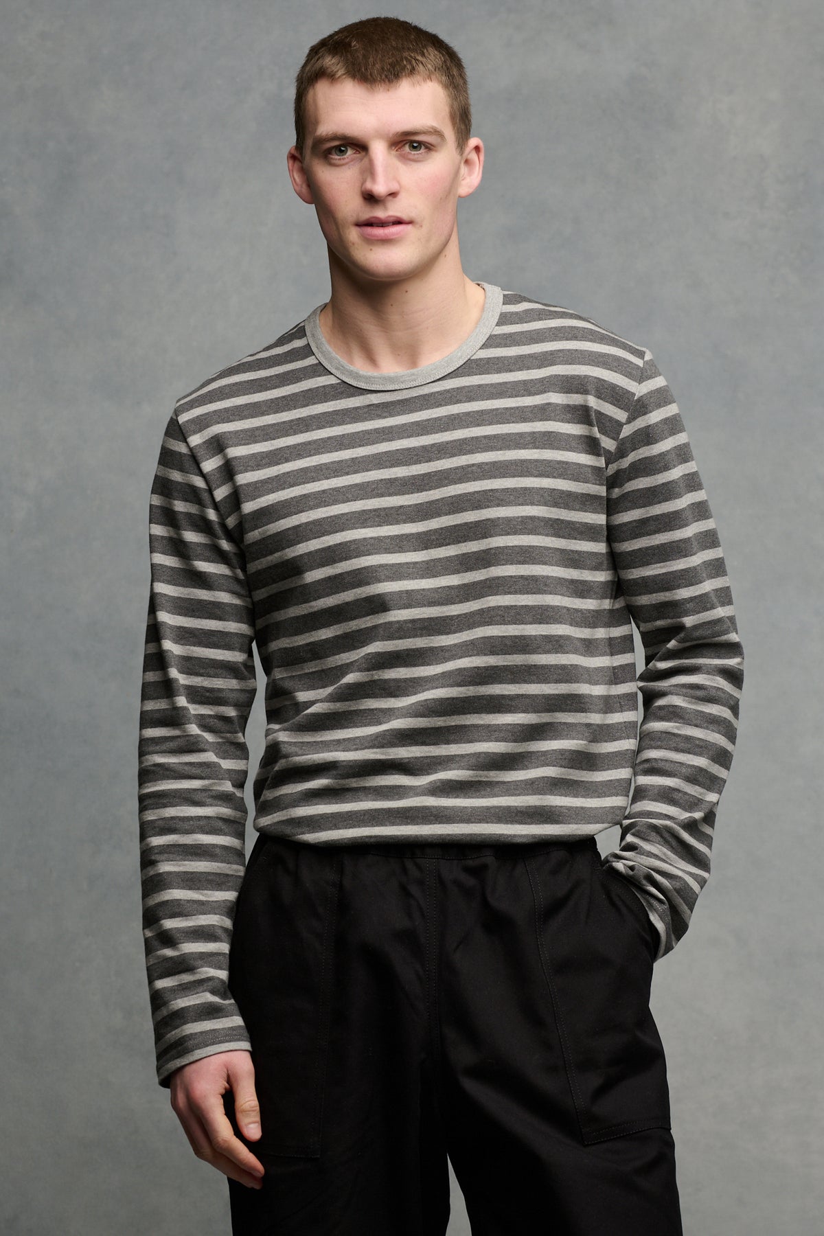 Thigh up image of the front of brunet male wearing charcoal marl/ grey marl tucked into cameraman pants in black 