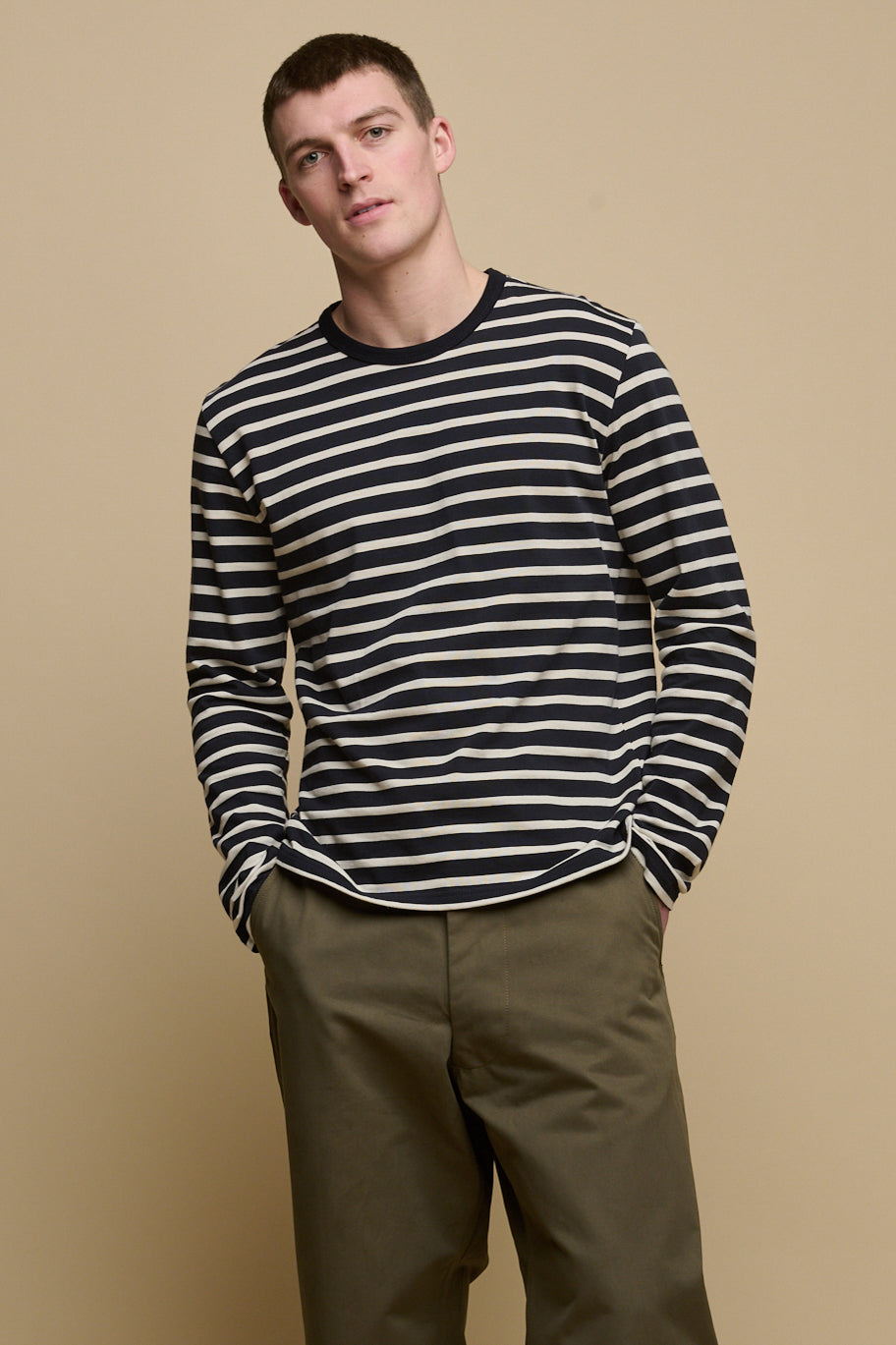 Thigh up image of male with shaved brunet hair wearing Breton in Navy ecru with his hands in front pockets of field trousers in olive