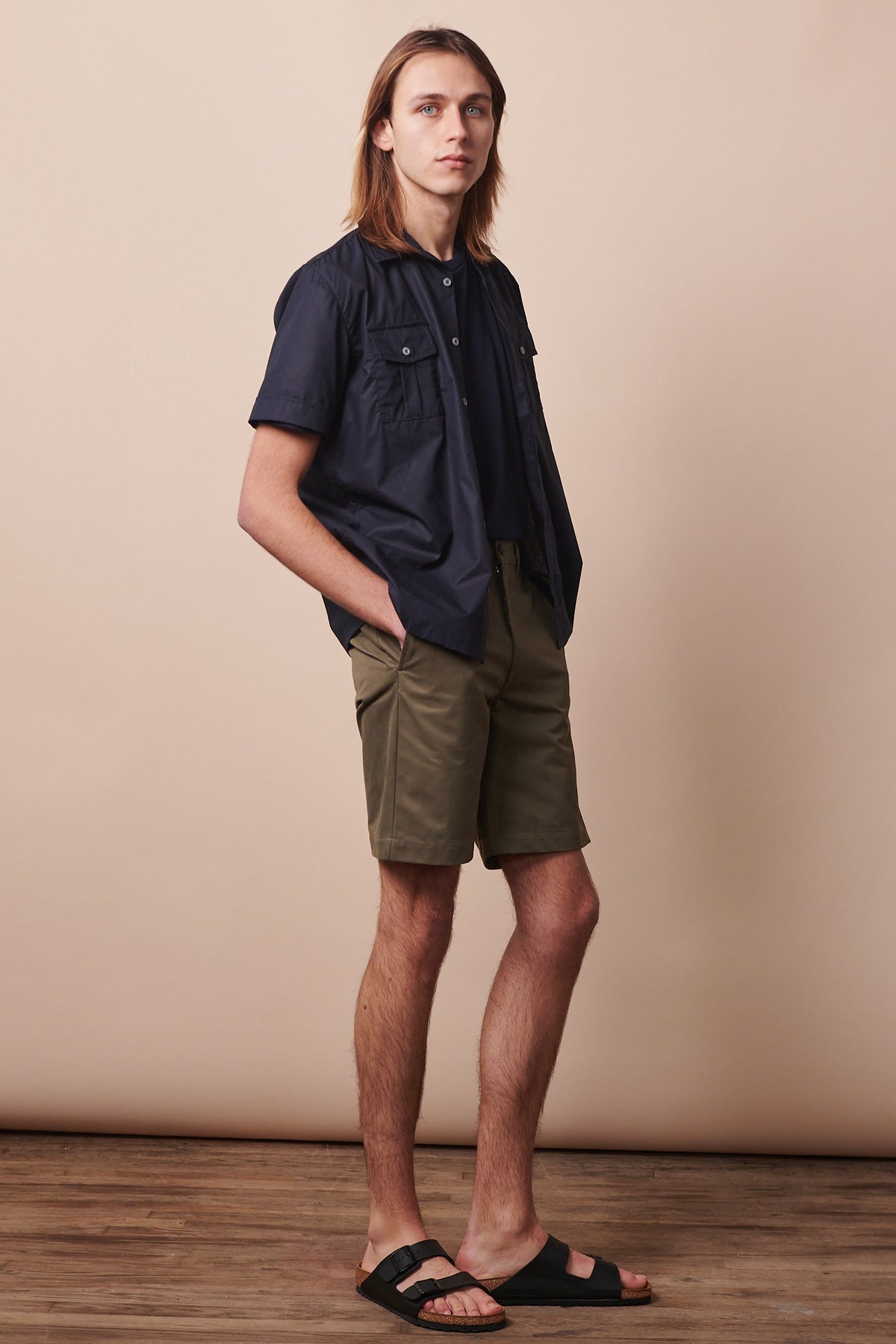 Full body image of the side of male wearing classic shorts in olive styled with unbuttoned military shirt in navy 
