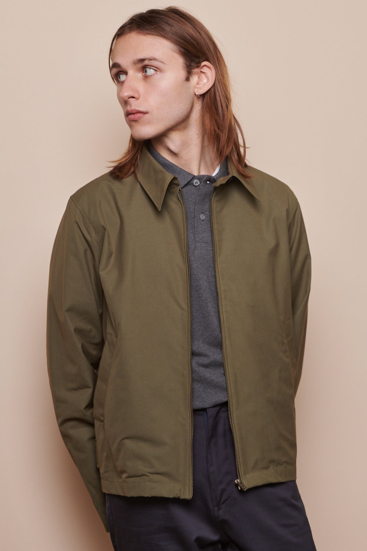 Thigh up image of male looking to the side wearing collared Harrington jacket in olive over polo shirt in grey
