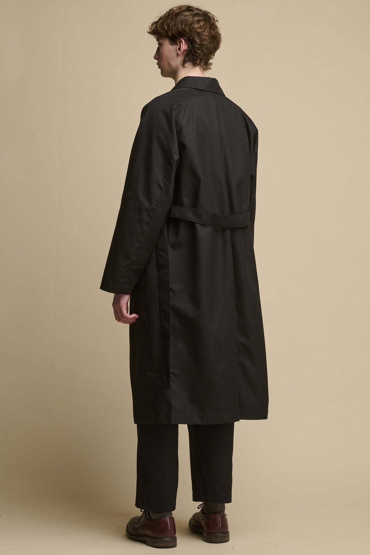 the back of male wearing Men's Frank Raglan Belted Raincoat in black. Belt held unfastened with two back belt loops 