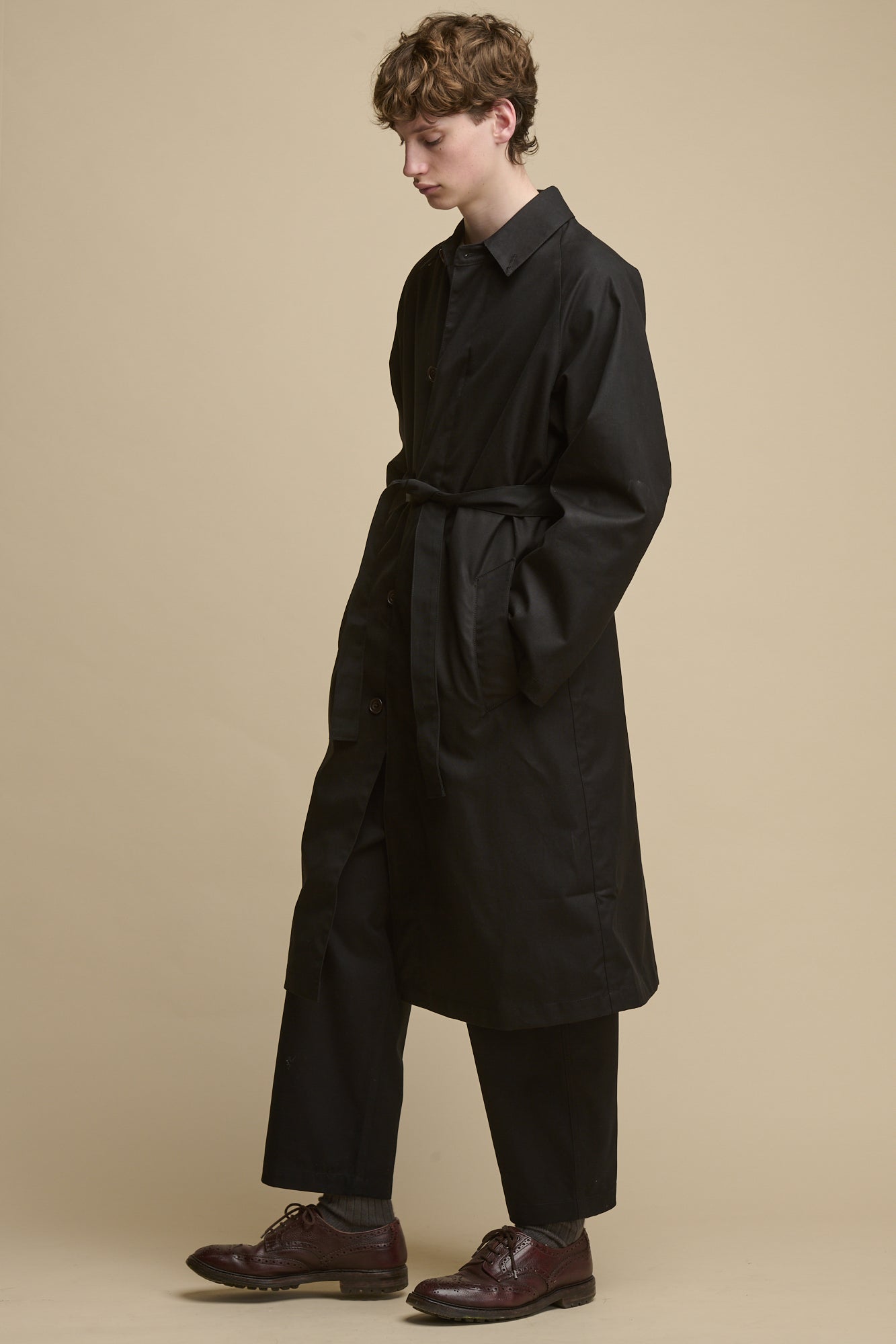 White male with orban hair wearing men's Frank Belted Raglan Raincoat in black, unbuttoned worn over stone raglan sweatshirt and black cameraman pants