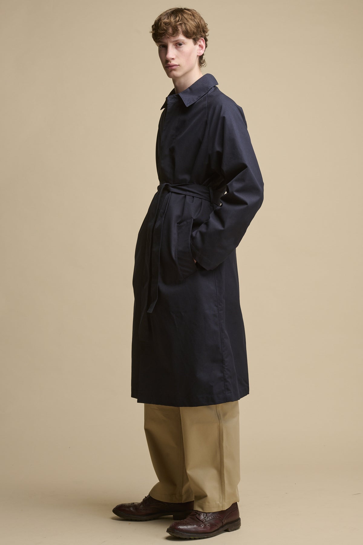 Full body image of male stood at an angle wearing Frank Raglan Belted Raincoat in navy with the belt fastened and his hands in the front two pockets.