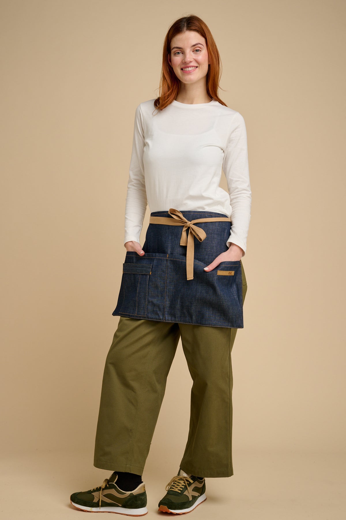 Woman wearing a white t shirt, denim apron, and green pants on a beige background.