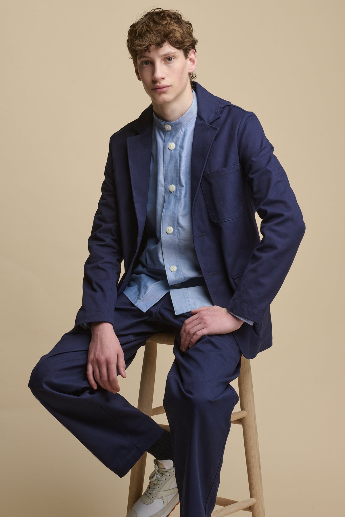 Full body image of male sat on stool wearing George Lightweight Collarless Overshirt in light blue paired with James Single Breasted Blazer in navy with cameraman pants in Navy