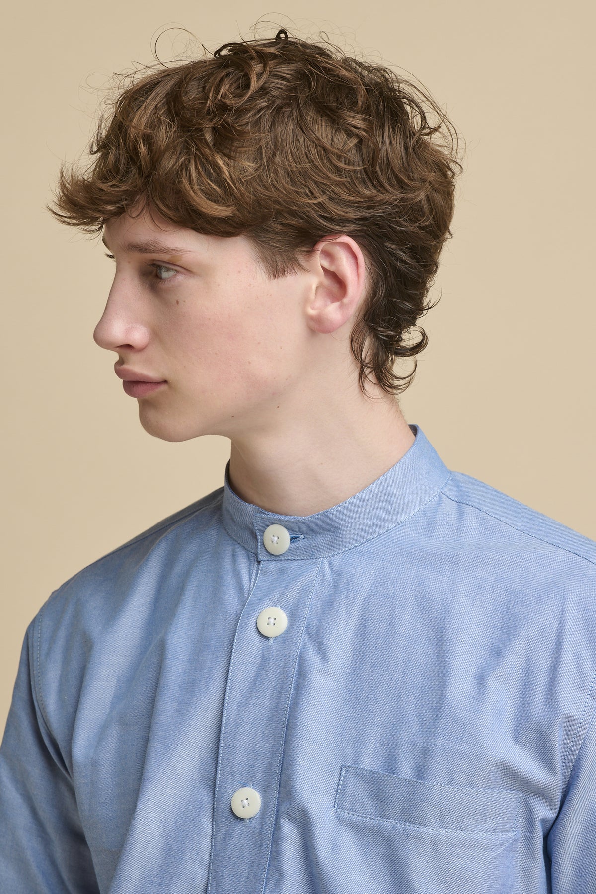 Portrait of male looking to the side wearing George Lightweight Collarless Overshirt in light blue with ecru white buttons