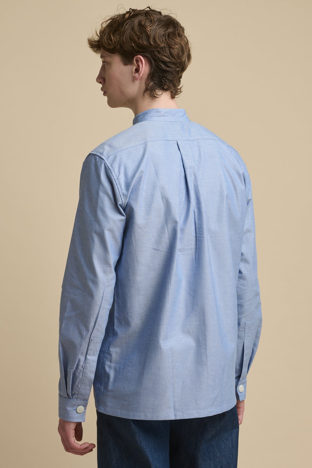 The back of male wearing George Lightweight Collarless Overshirt in light blue with back pleat and button cuff