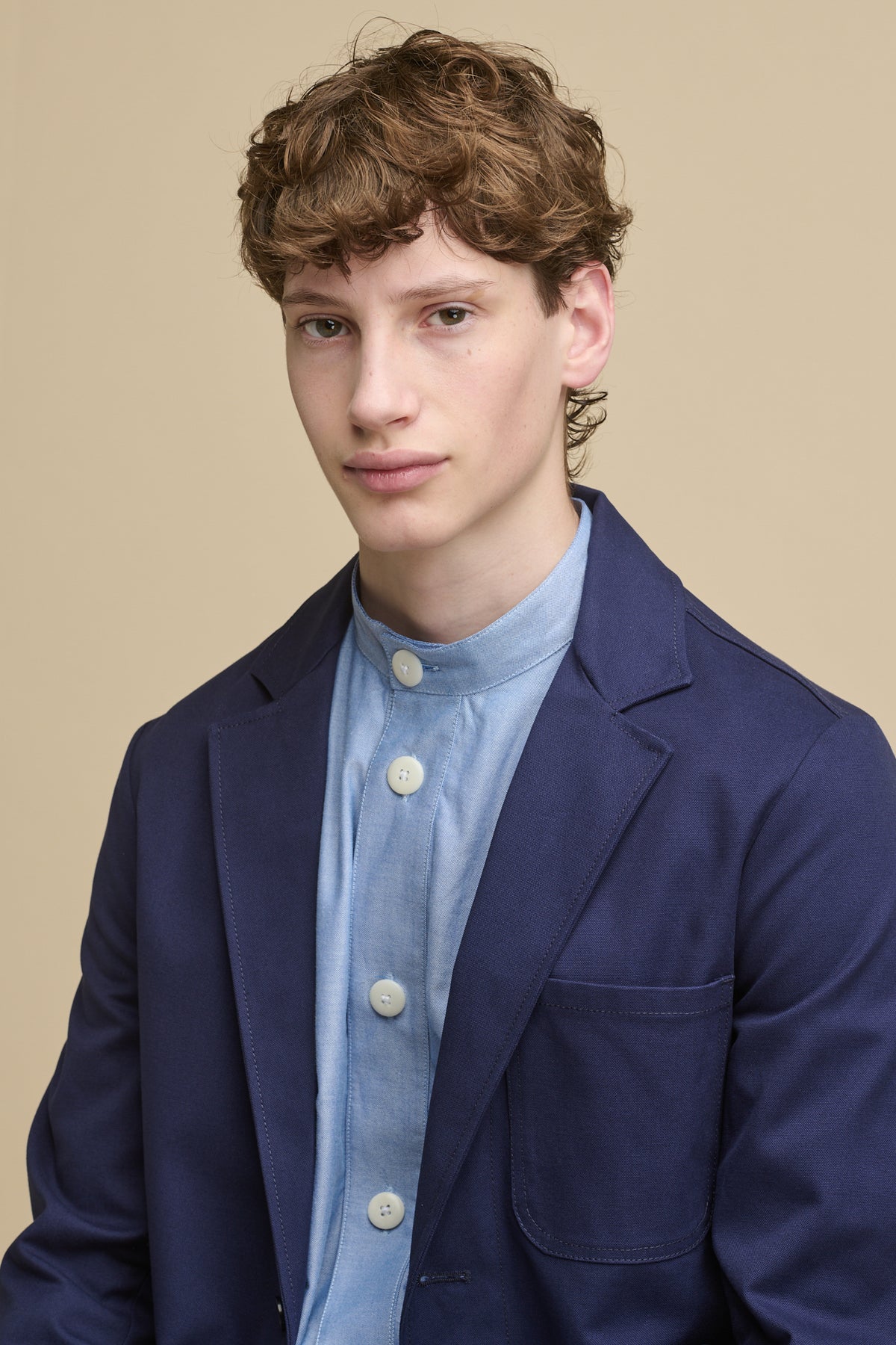 Portrait of male wearing George Lightweight Collarless Overshirt in Light Blue with large ecru white buttons worn underneath James Single Breasted Blazer in navy