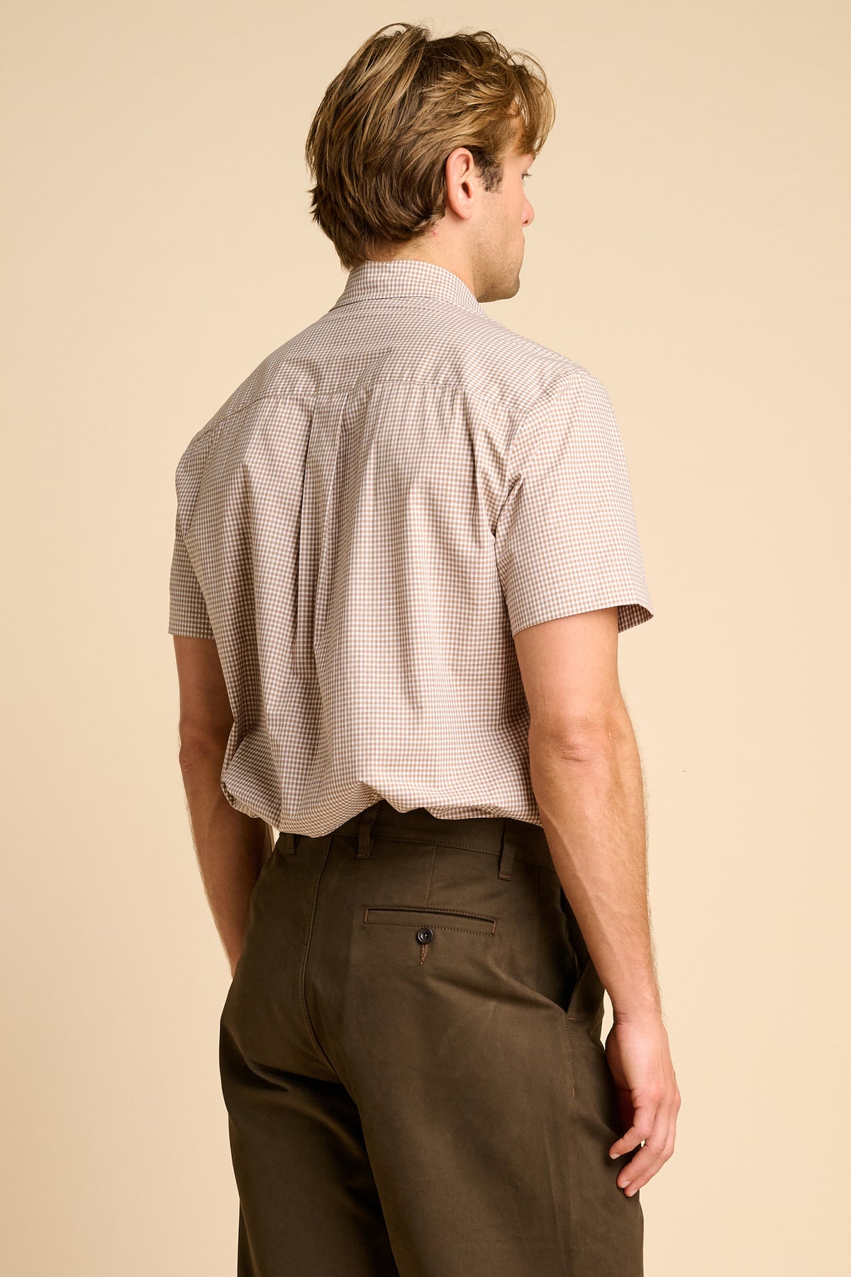 A man wearing beige short-sleeve button-up shirt and dark brown trousers, standing against a beige background.