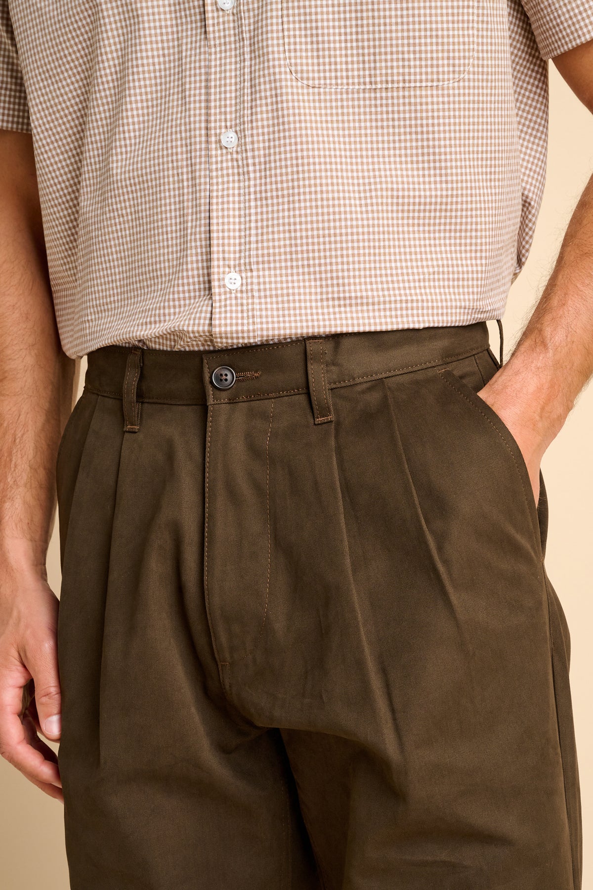 Close-up of a person wearing dark brown pleated trousers and a beige and white checkered short sleeve button up shirt, with one hand in a pocket.