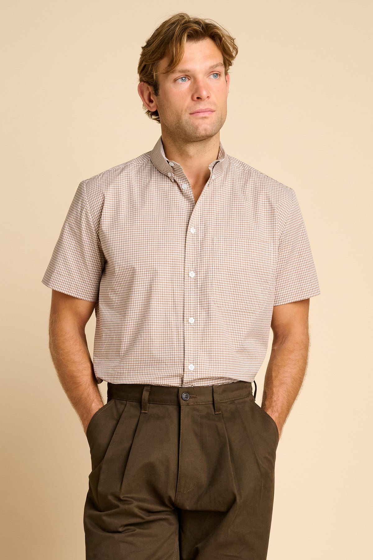 A man wearing a short sleeve button down shirt paired with dark brown pleated trousers, against a beige background.