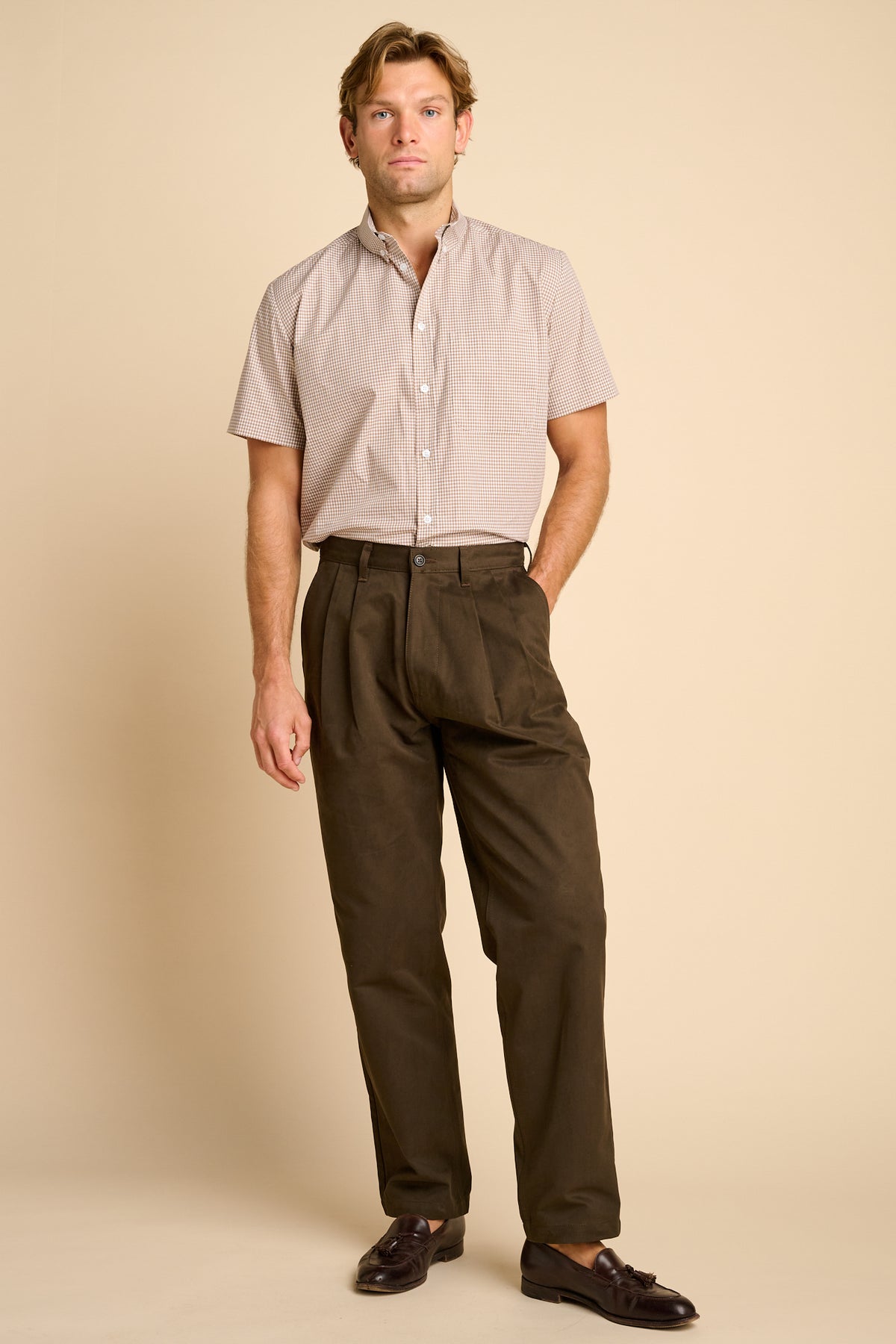A man wearing beige short-sleeve button-up shirt and dark brown trousers, standing against a beige background.