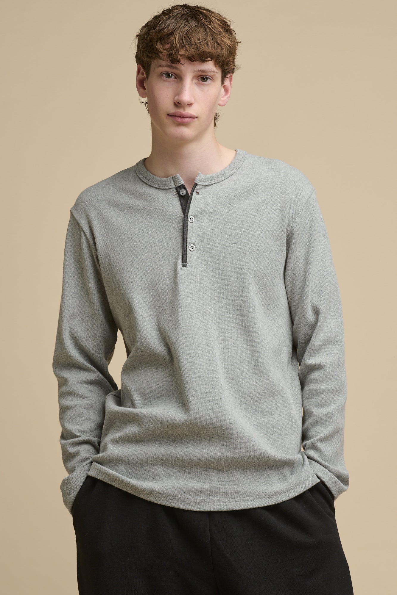 Thigh up front of male wearing long sleeve Henley top in grey marl with his hands in the pockets of black sweatpants