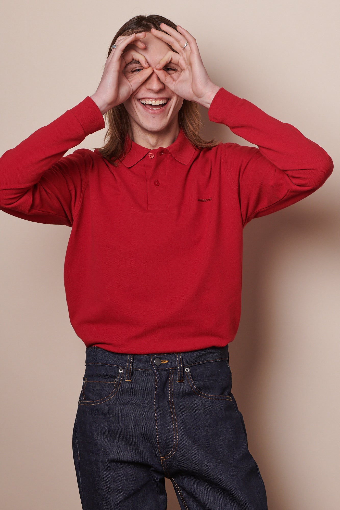 Thigh up image of white male with shoulder length brown hair wearing long sleeve t shirt in crimson with arms held behind back 