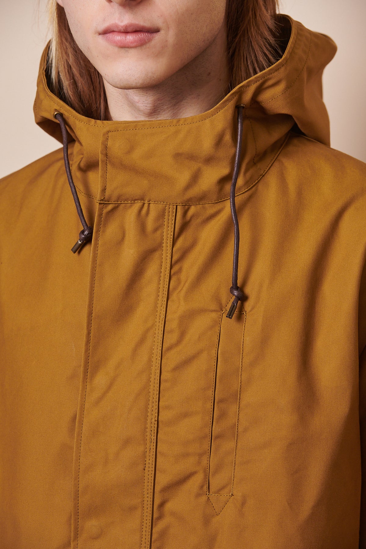 Close up image of male wearing zipped up mountain parka in tan, brown draw cord through hood.