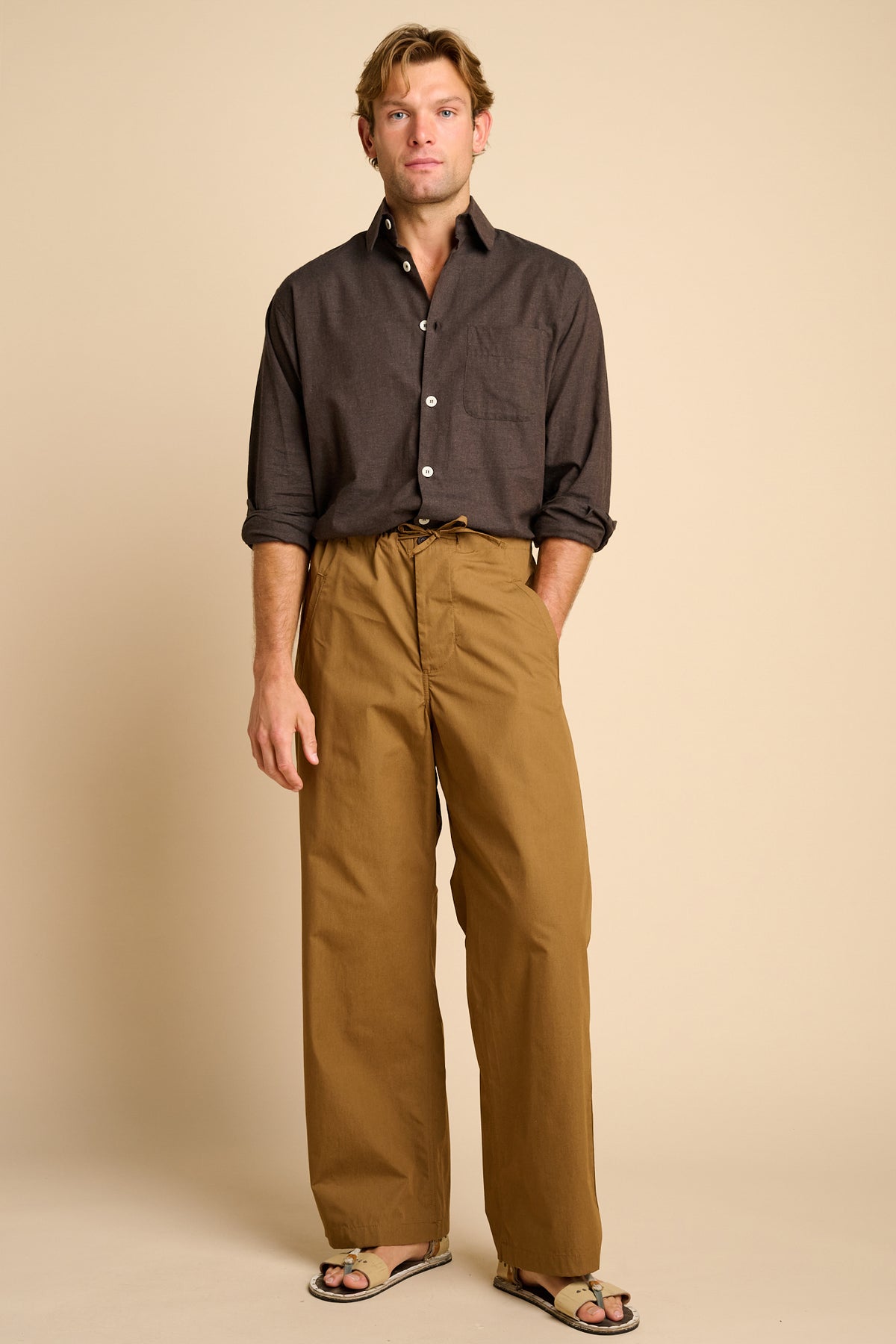 Man wearing tan trousers with brown shirt  against beige background.