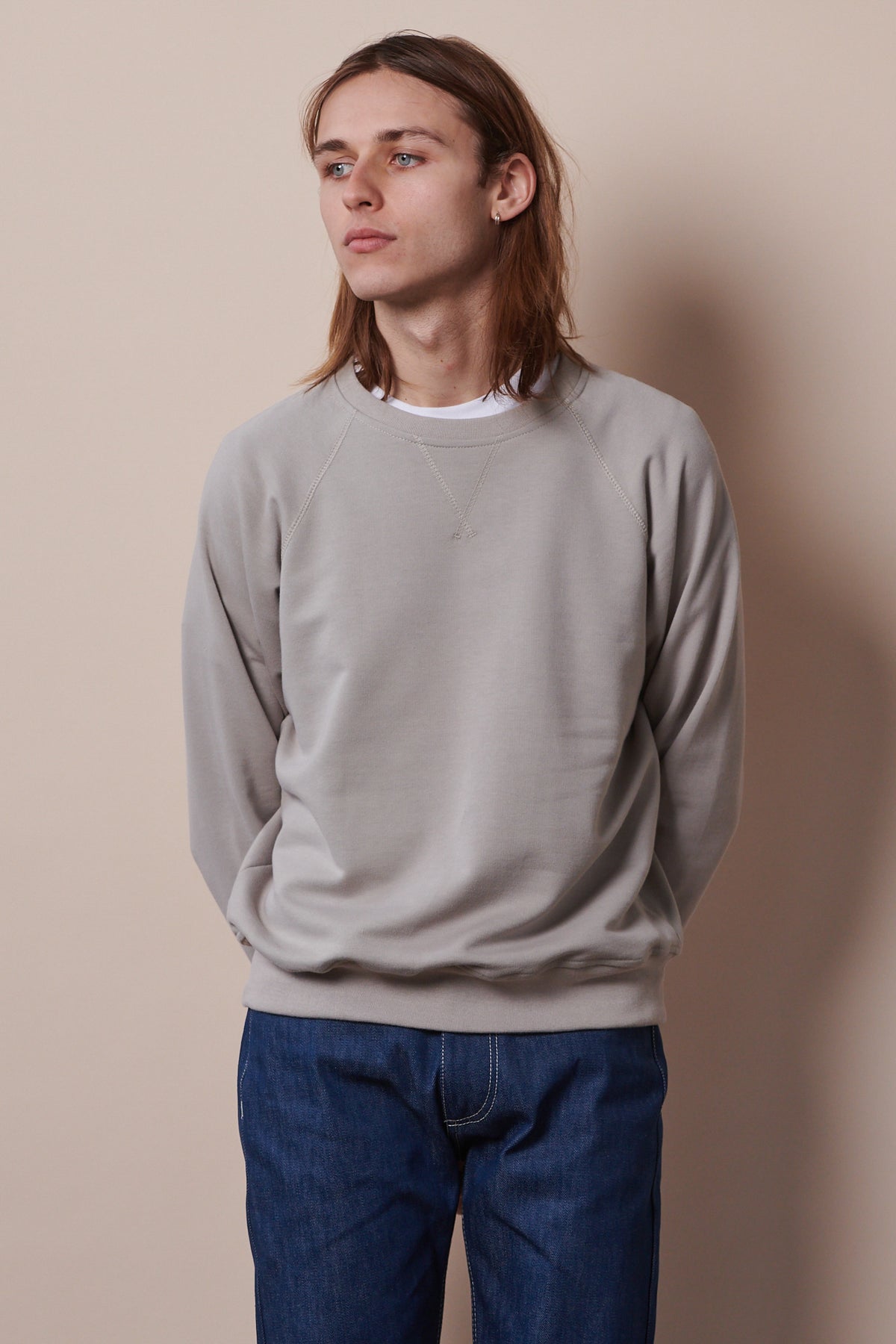 Thigh up image of white male with shoulder length brunet hair wearing raglan sweatshirt in stone paired with straight cut jeans in blue