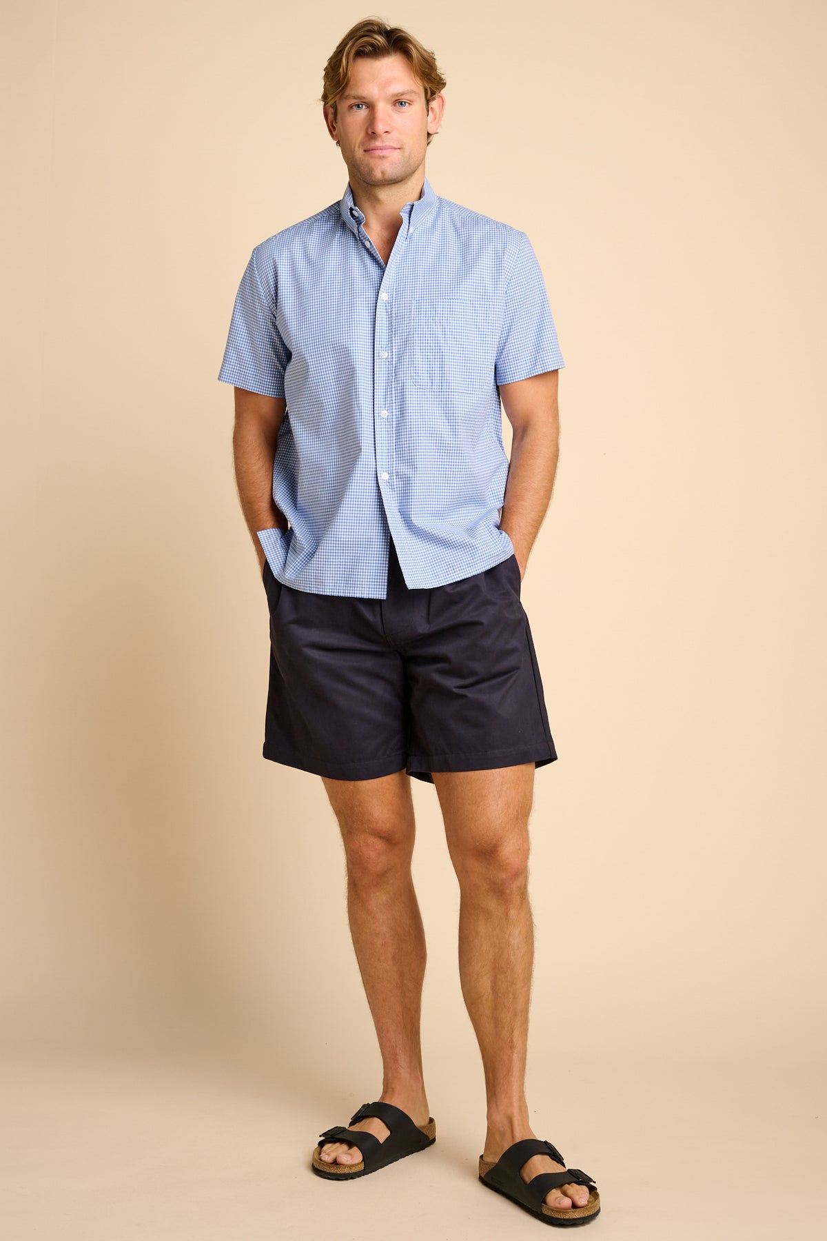 Man wearing a light blue checkered shirt and dark shorts on a beige background.