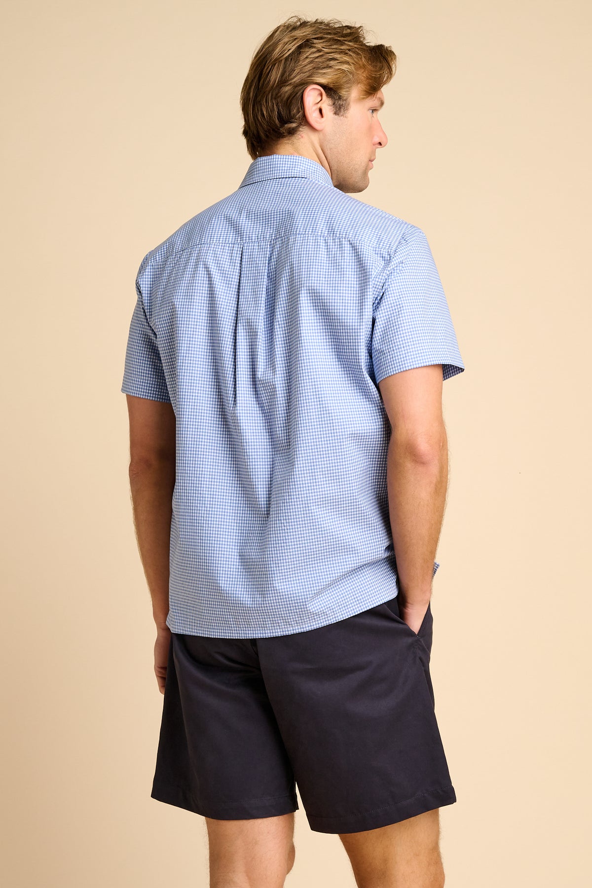 Man wearing a light blue checkered shirt and dark shorts on a beige background.