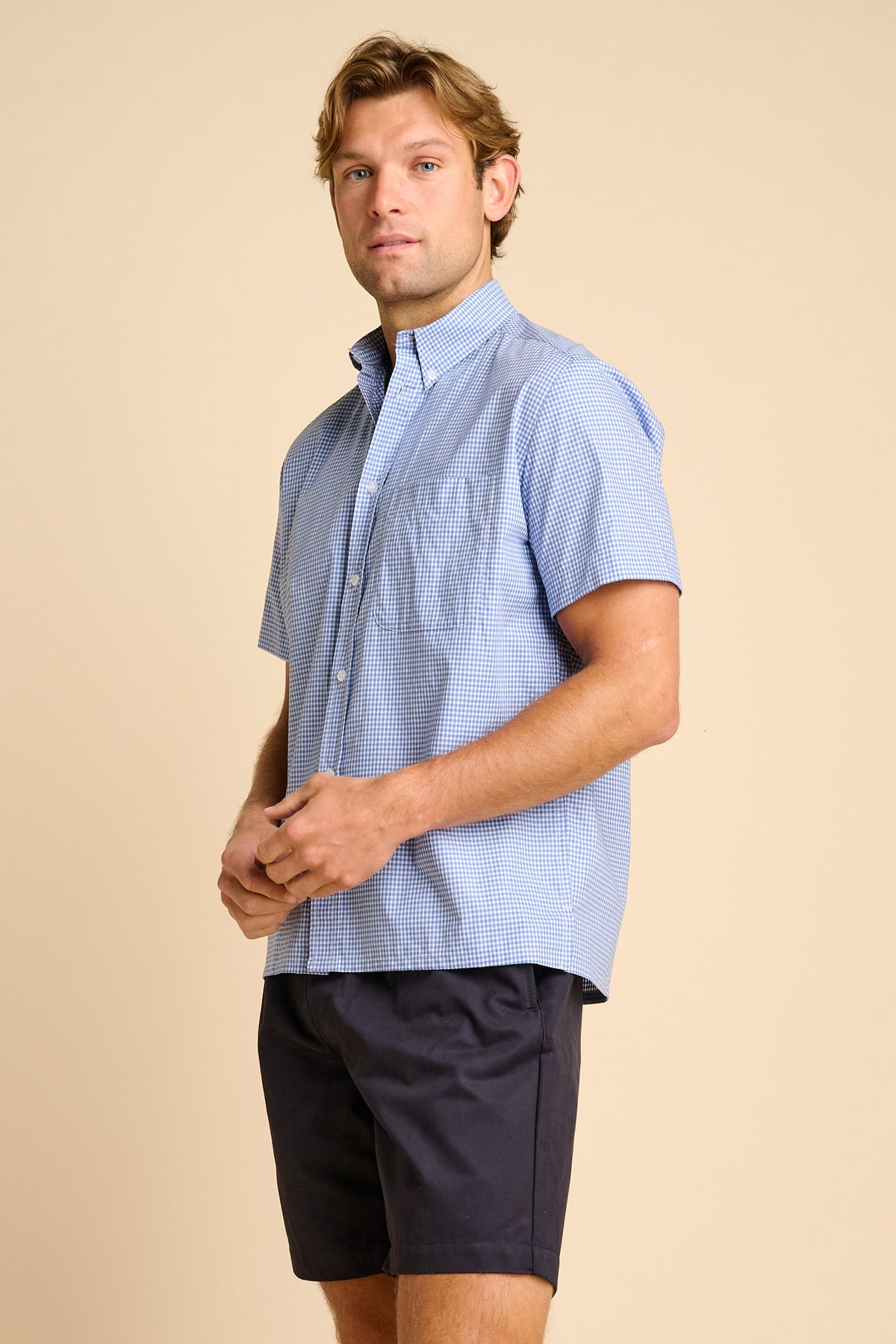 Man wearing a light blue checkered shirt and dark shorts on a beige background.