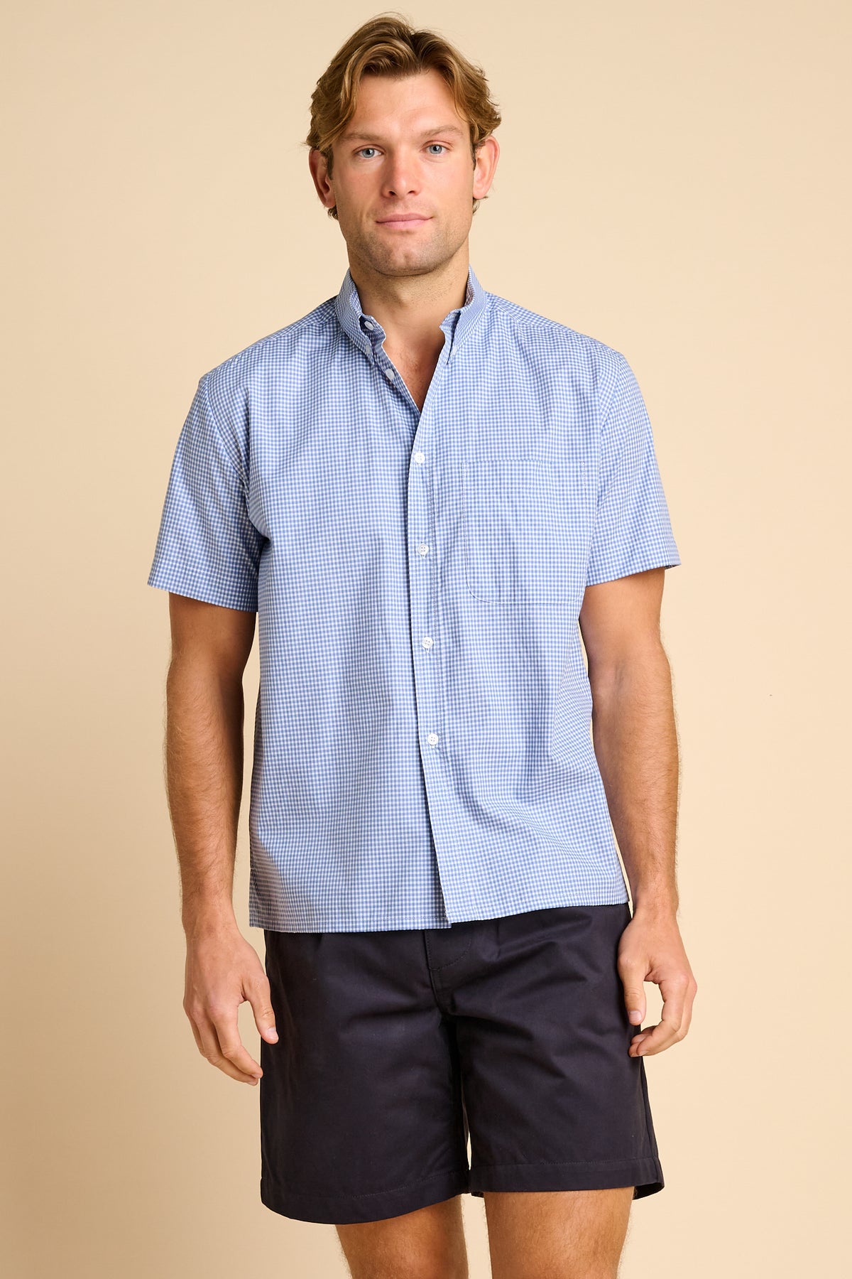 Man wearing a light blue checkered shirt and dark shorts on a beige background.