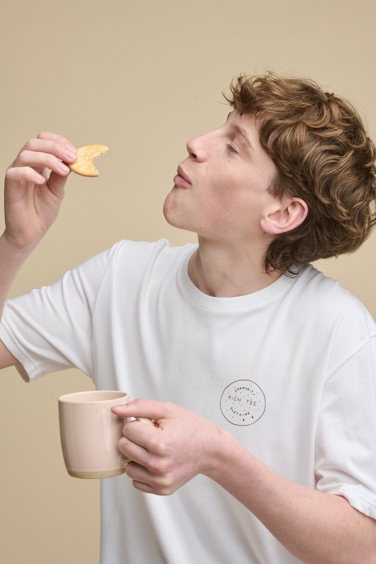 Male holding a half eaten rich tea biscuit up in one hand with a mug in the other, wearing short sleeve graphic t shirt with rich tee print on
