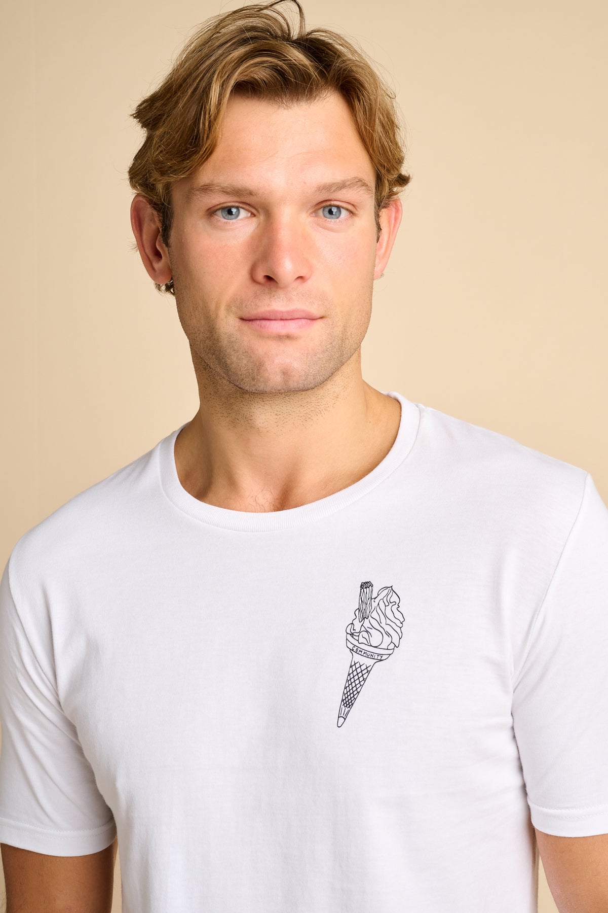 A man wearing a white crew neck t-shirt with a small black line art graphic of an ice cream cone on the left chest, set against a solid beige background.
