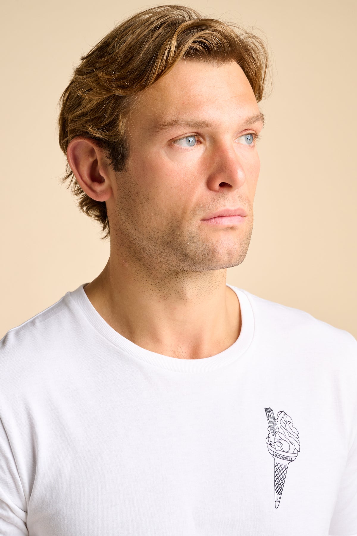 A close-up of a man wearing a white crew-neck t-shirt with a small black line art graphic of an ice cream cone on the left chest, set against a solid beige background.