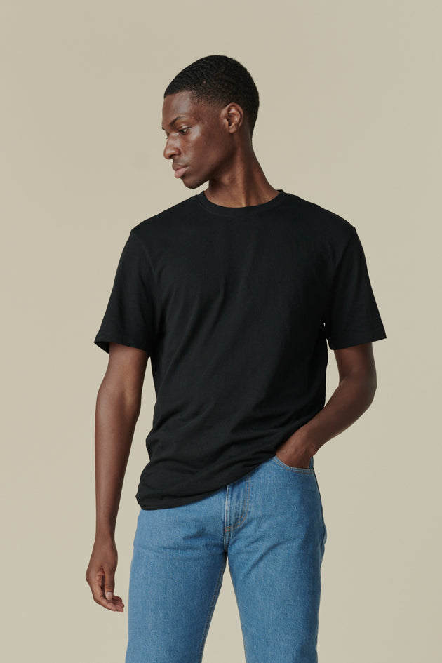 Thigh up image of male from the front looking to the side away from the camera, wearing short sleeve t shirt plastic free in black with straight cut selvedge jeans in fade