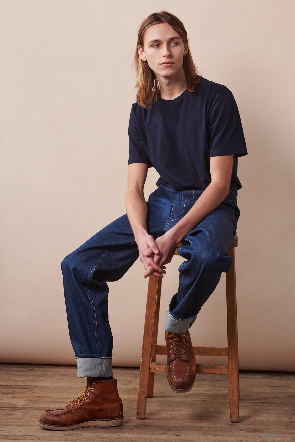 Full body image of male sat on stool with arms leaning on legs wearing short sleeve t shirt in navy paired with chore jeans in blue