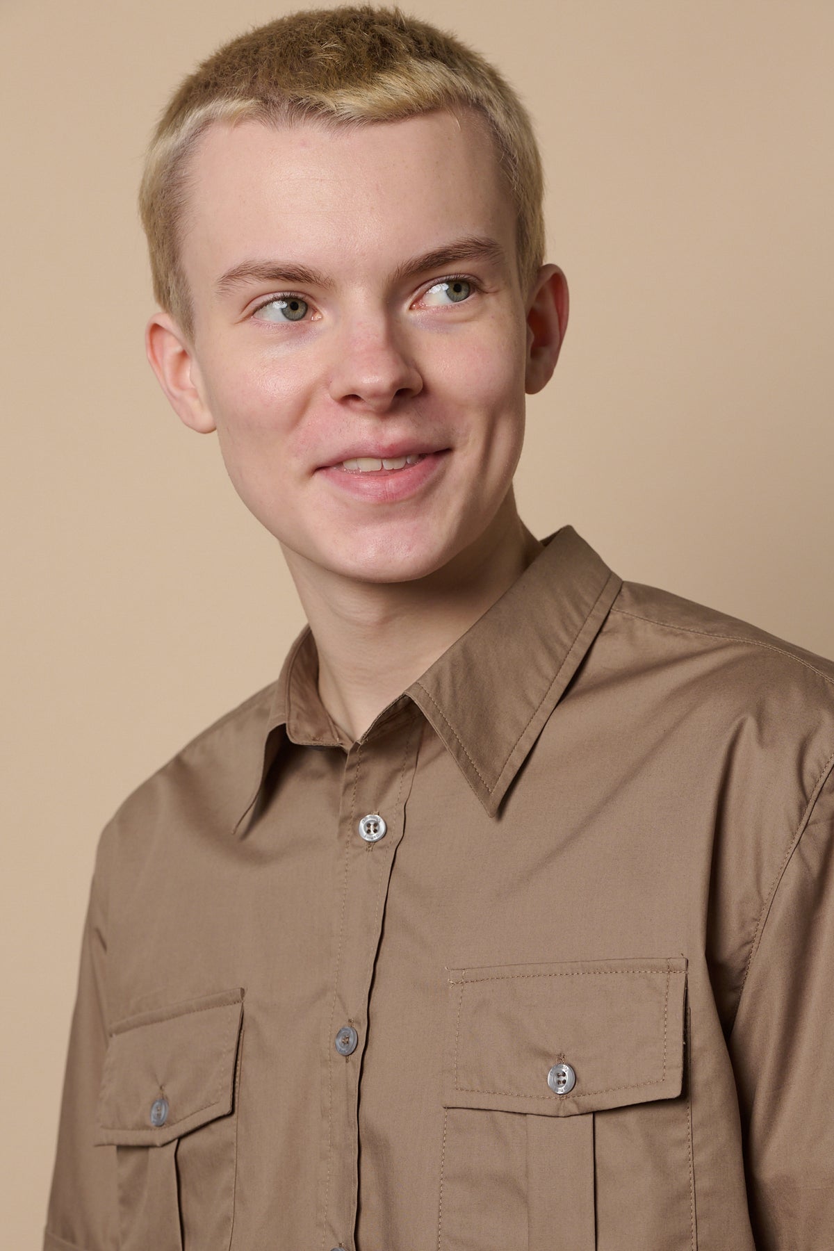 Portrait of smirking male with shaved, short blond hair wearing Tom short sleeve military shirt with top button undone.  