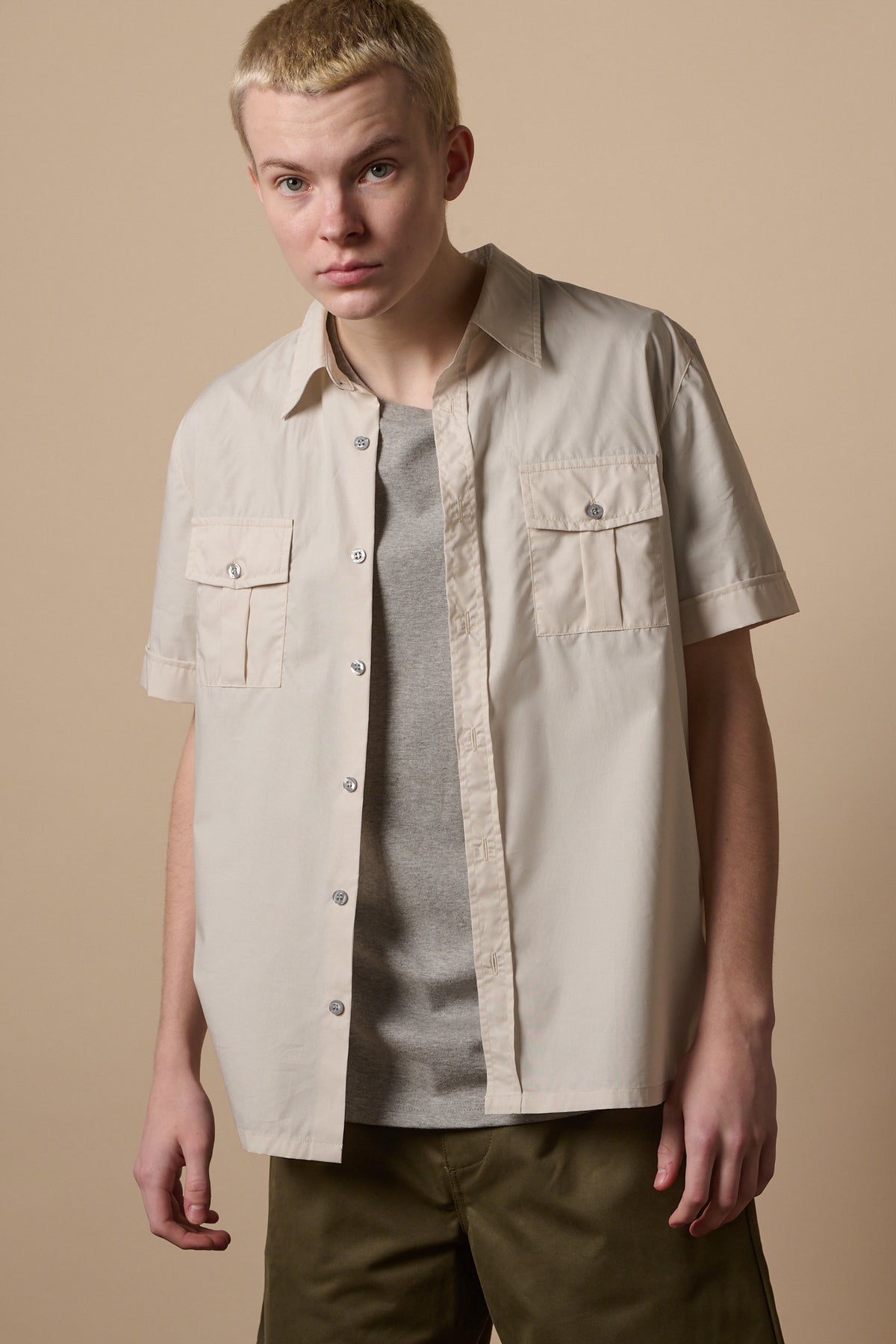 thigh up image of male with short shaved blond hair wearing Tom short sleeve military shirt in stone unbuttoned over short sleeve crew neck t shirt in stone