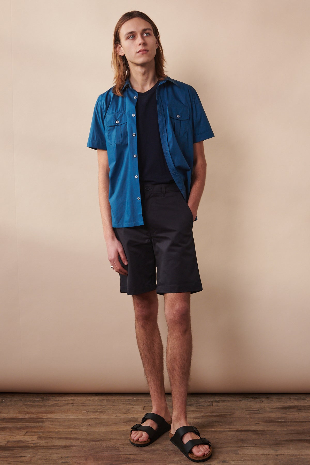 Full body image of male wearing unbuttoned Tom short sleeve military shirt in light blue over short sleeve t shirt in navy paired with navy chino shorts and Birkenstock sandals