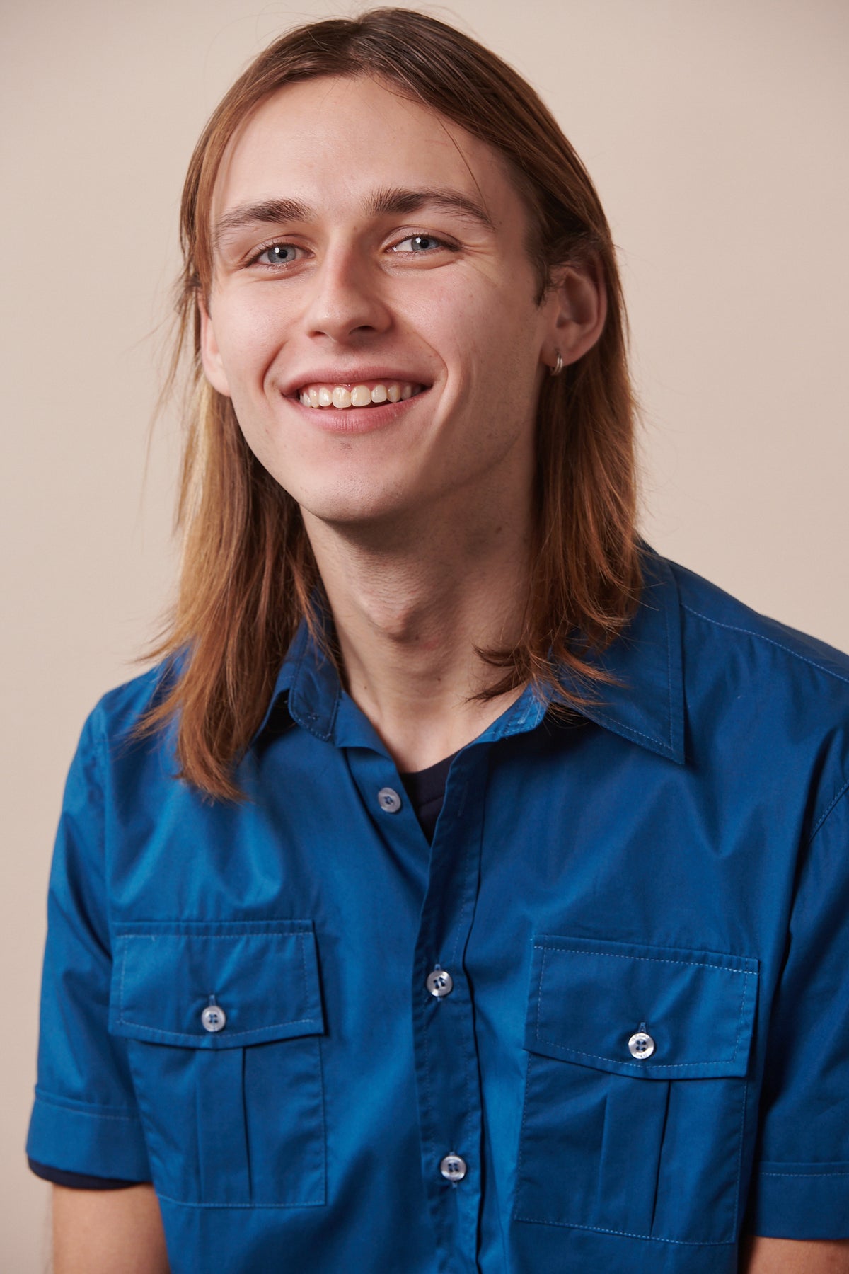 Portrait of smiley male with shoulder length brown hair wearing Tom short sleeve military shirt in light blue buttoned over short sleeve t shirt in navy