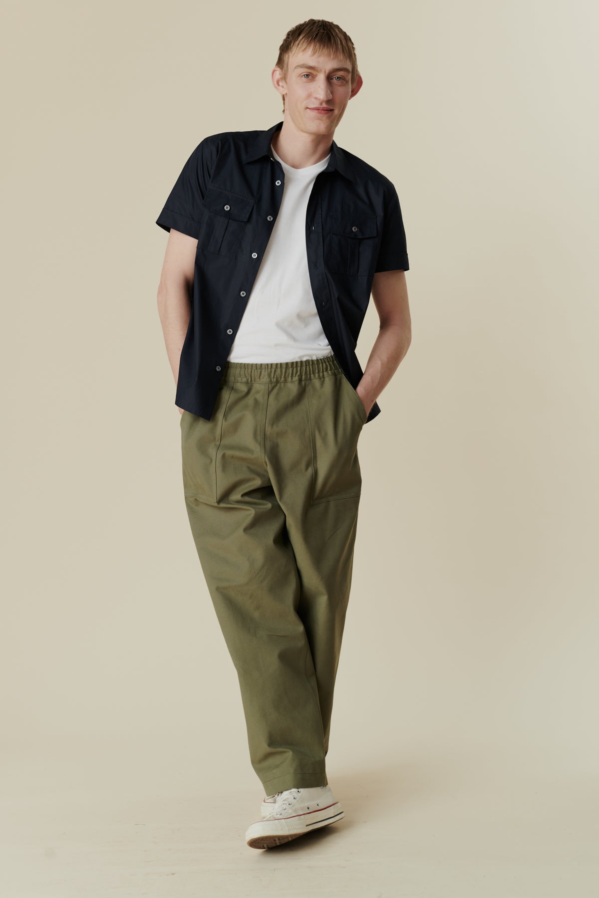 Full body image of blonde male wearing tom short sleeve military shirt unburttoned over short sleeve white crew neck t shirt in white, paired with olive cameraman pants
