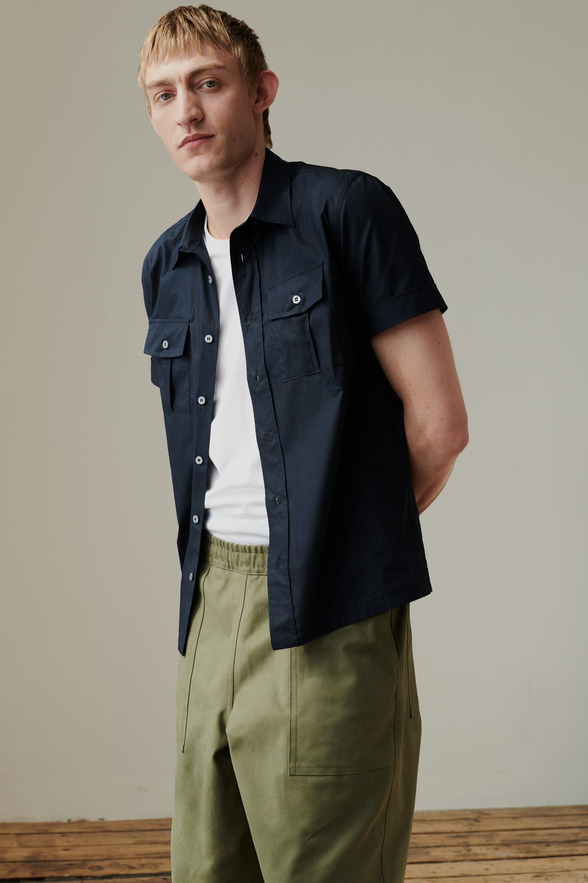 Knee up image of white male holding arms behind his back wearing tom short sleeve military shirt unbuttoned over white t shirt