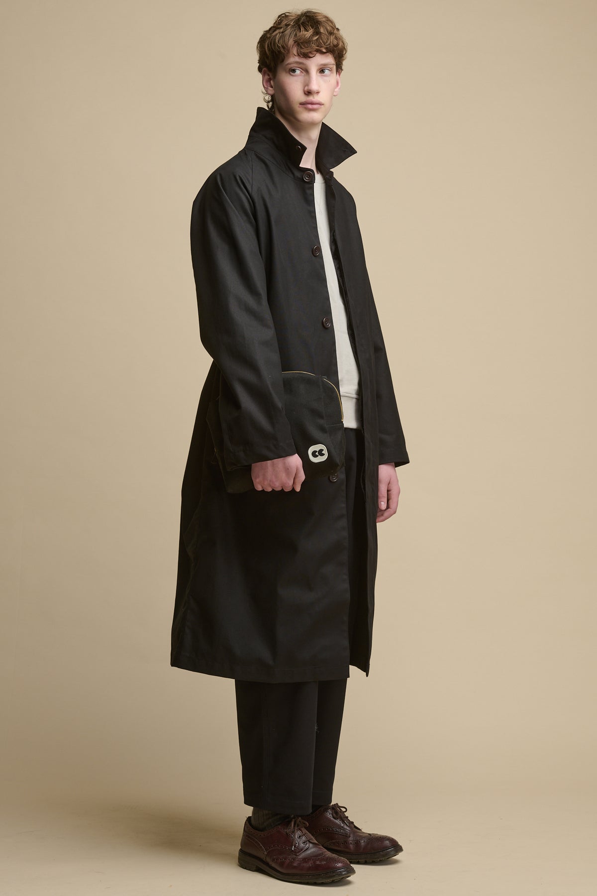 Full body image of male wearing Frank raglan belted raincoat in black holding pouch in black with CC patch logo