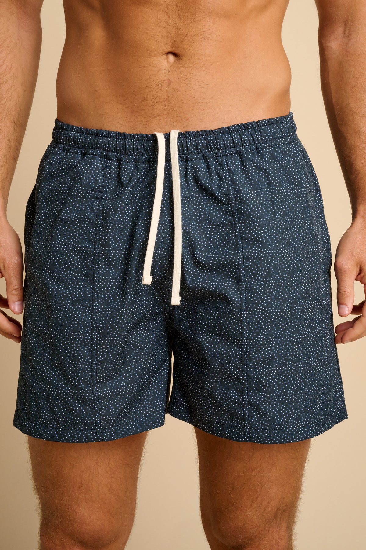 Navy polka dot swim shorts with a white drawstring on a beige background.