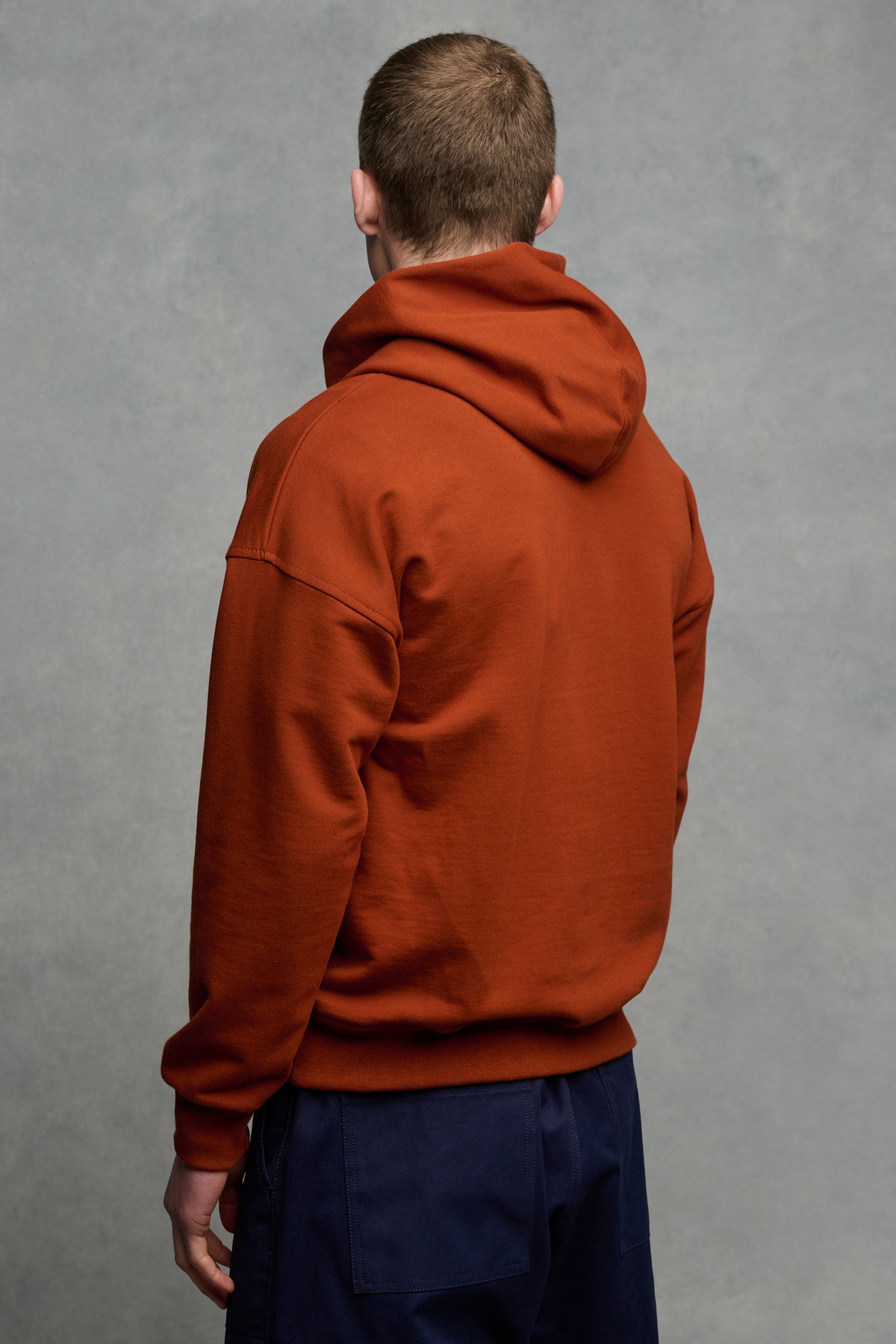 The back of brunet male wearing hooded sweatshirt  in cinnamon paired with navy cameraman pant