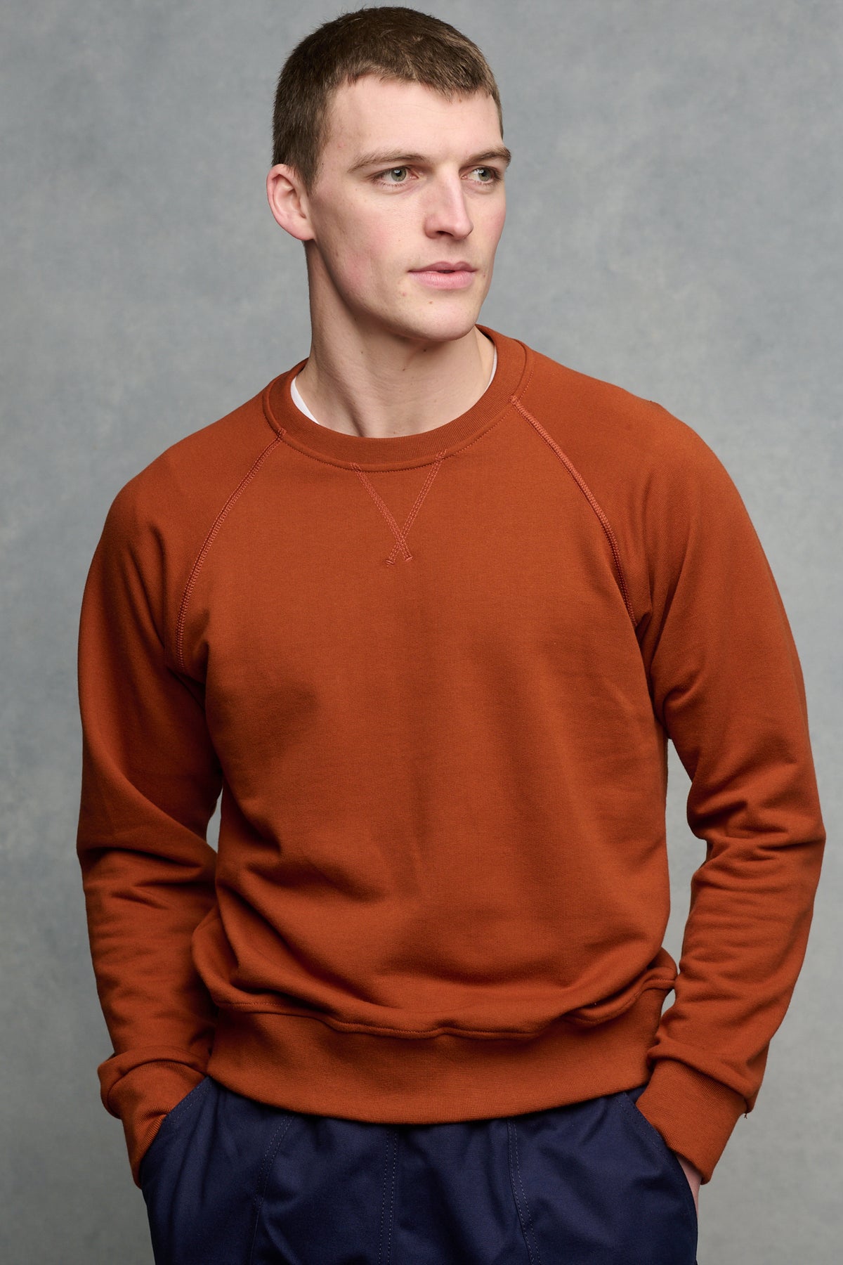 Thigh up image of brunet male wearing raglan sweatshirt in cinnamon worn over crew neck short sleeve t shirt in white, paired with cameraman pant in navy