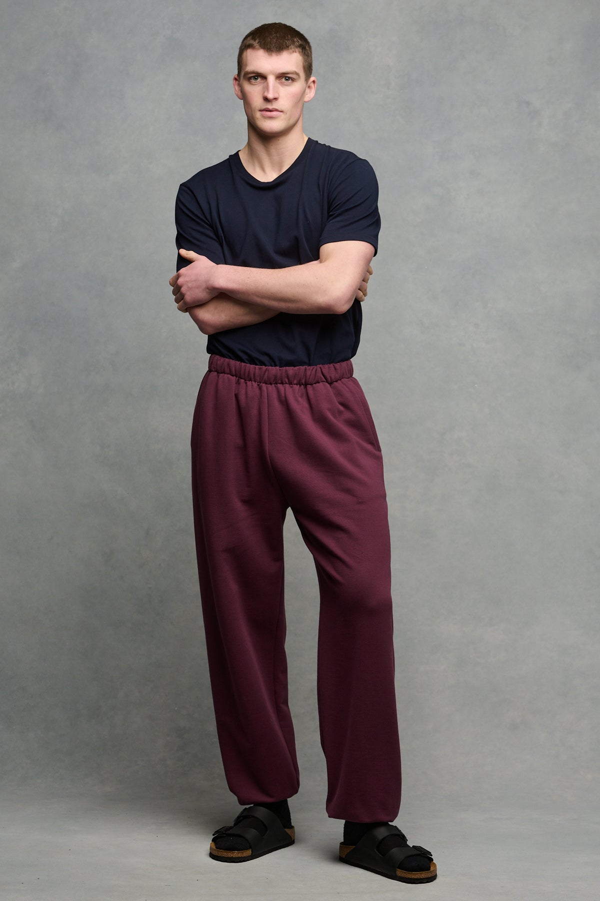Full body image of brunet male wearing sweatpants in plum with crew neck t shirt in navy tucked into sweatpants 